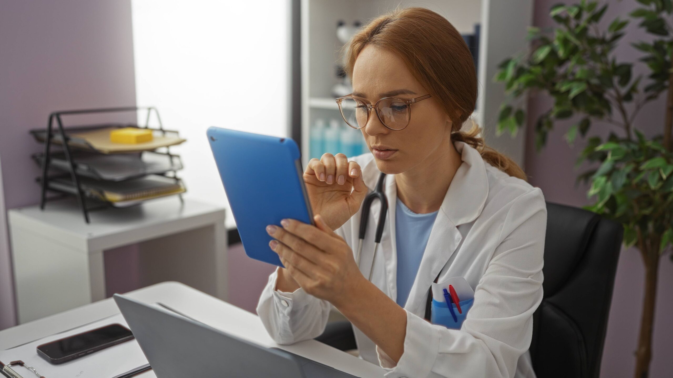 Female doctor with glasses reviewing patient records on blue tablet in modern medical office
