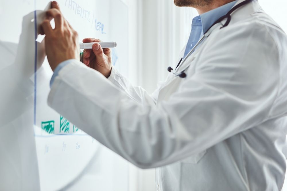 A doctor with a stethoscope wearing a white lab coat writes on a whiteboard with a marker.