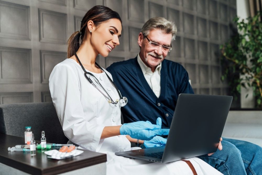 A female doctor in a white coat shows an elderly male patient something on a laptop.